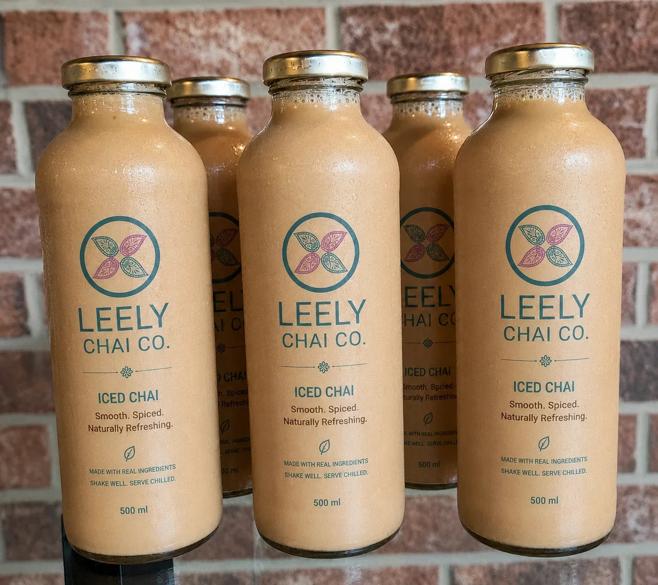 Five glass bottles of LEELY Chai Co. Iced Chai with branded labels and gold caps displayed against a rustic brick wall.