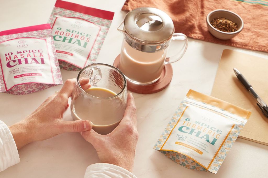 Hands holding a glass of freshly brewed chai beside a French press and Leely Chai Co. blends, including Masala Chai, Rooibos Chai, and Turmeric Chai, on a cozy tabletop.