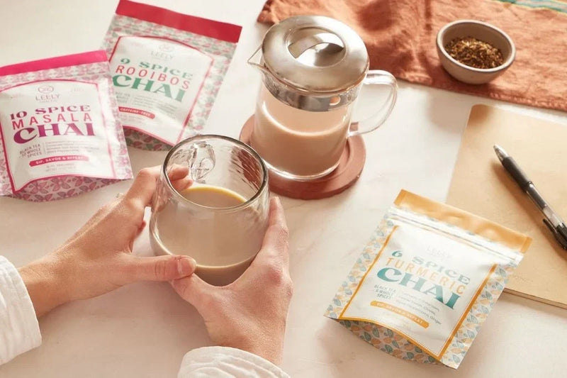 Hands holding a warm cup of chai beside Leely Masala, Rooibos, and Turmeric Chai blends on a bright morning desk.