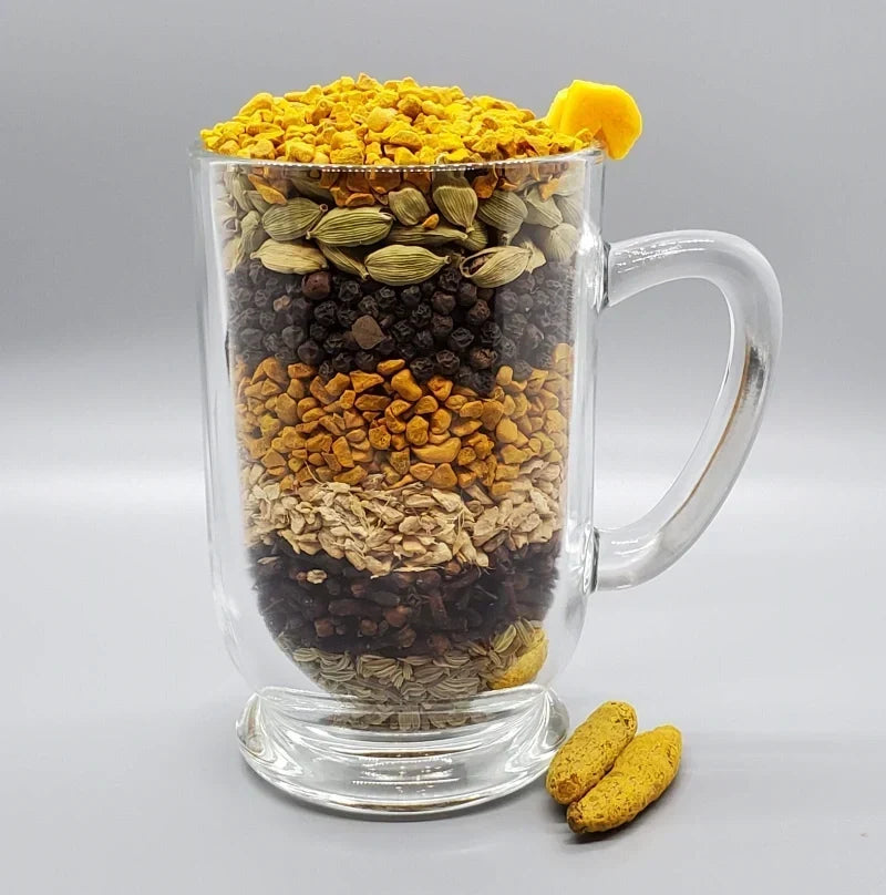 Glass mug layered with turmeric, ginger, cardamom, black pepper, fennel, cloves, and other whole chai spices.