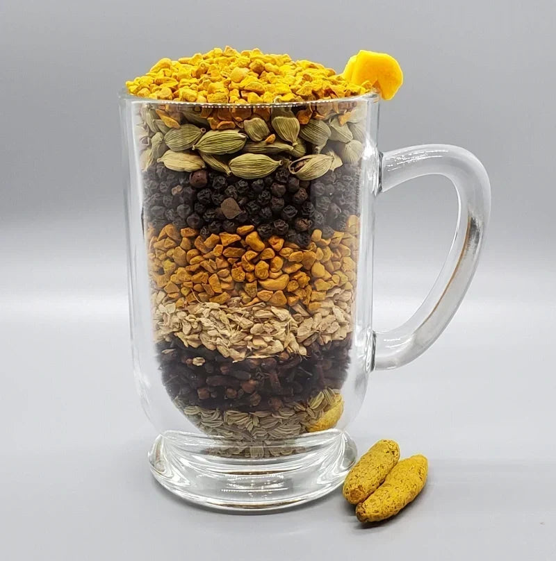 Glass mug layered with turmeric, ginger, cardamom, black pepper, fennel, cloves, and other whole chai spices.
